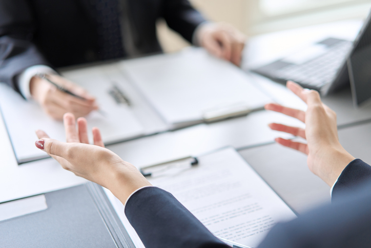 Two people in business attire sitting at a desk with documents and a laptop, engaged in a discussion. One person gestures with open hands while the other holds a pen, appearing to explain or negotiate.