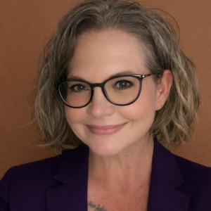 A woman with short, wavy gray hair and glasses smiles warmly. She is wearing a purple blazer and is posed in front of a plain brown background.