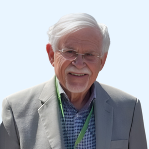 An older man with white hair and glasses smiles, wearing a light gray blazer, a checkered shirt, and a green lanyard, standing against a light blue background.