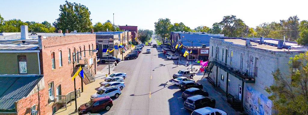 Aerial view of a small town main street lined with parked cars, brick buildings, and Ukrainian flags hanging from lamp posts on both sides. Trees and clear blue sky are visible in the background.
